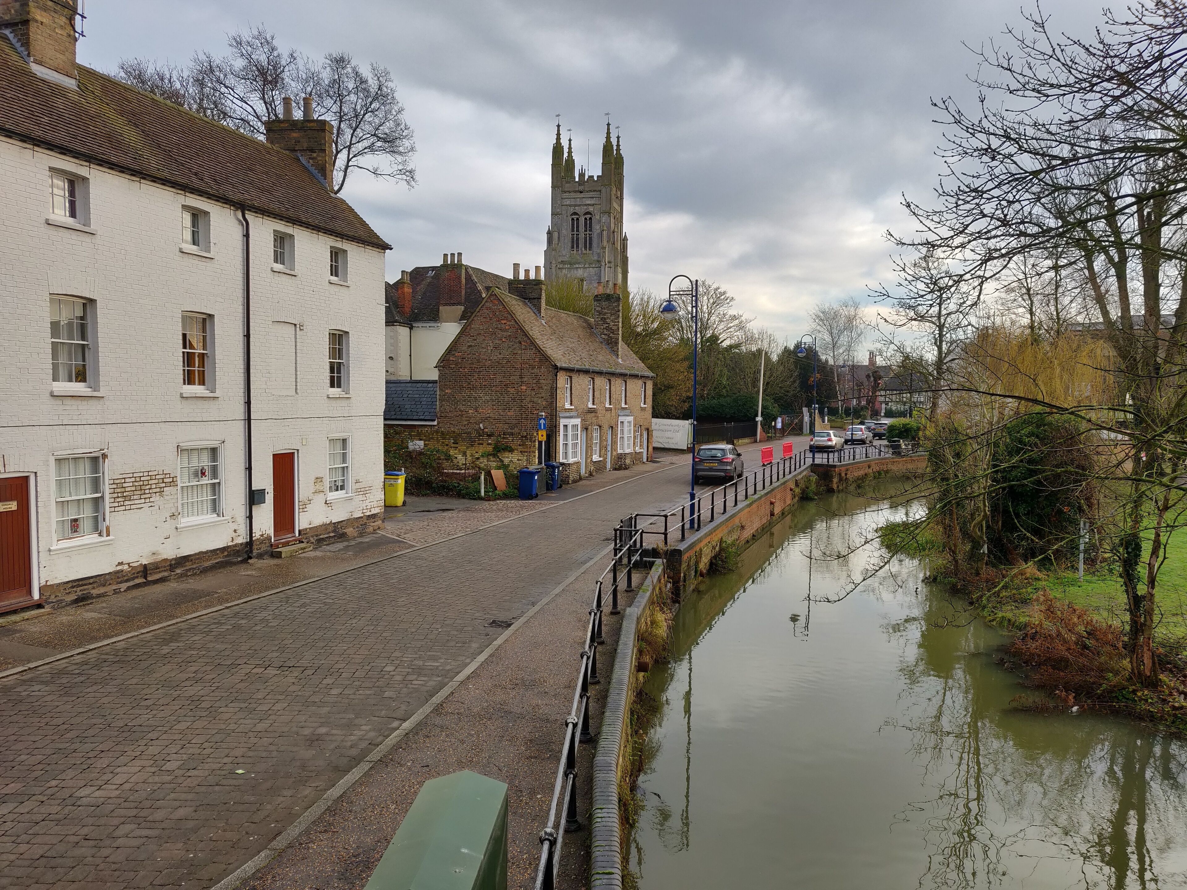 A cobblestone street lined with historic buildings, a canal, and a church tower in the background.