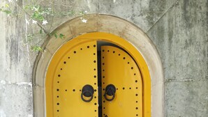 Ein gelbes, rundbogenförmiges Portal mit zwei runden Metallgriffen, in eine Steinmauer eingelassen.