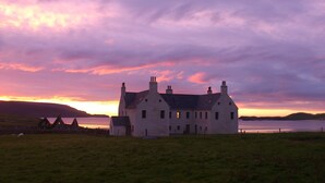 A large, white building with multiple chimneys, situated on a grassy field with a body of water and hills in the background.