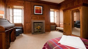 A room with wooden paneling, a fireplace with flowers, a bed with a plaid blanket, a piano, and a large mirror.