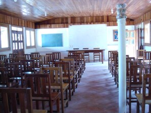 A classroom with wooden chairs and tables arranged in rows, a whiteboard, and a large window with blinds.