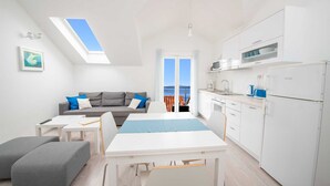 A modern, well-lit room with a dining table, a grey sofa with blue cushions, a grey ottoman, a kitchen area with white appliances, and a large window with a view of the sea. - Penthouse apartment Sea (Bol)