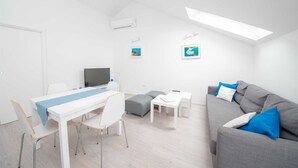 A modern, well-lit room with a white ceiling, a grey sofa with blue cushions, a white dining table with chairs, a grey ottoman, and a flat-screen TV. - Penthouse apartment Sea (Bol)