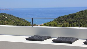 Ein Balkon mit drei schwarzen Sitzgelegenheiten, der auf eine Küstenlandschaft mit Bäumen und dem Meer blickt.