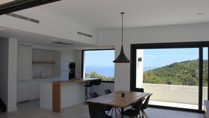 Eine moderne Küche mit einer großen Kücheninsel, einem Essbereich mit Holztisch und Stühlen sowie einem großen Fenster mit Blick auf das Meer.