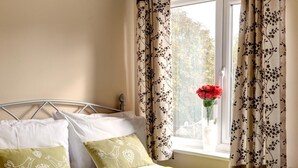 Una cama bien hecha con colcha verde, dos almohadas con diseños de pájaros y un florero con flores rojas en el alféizar.