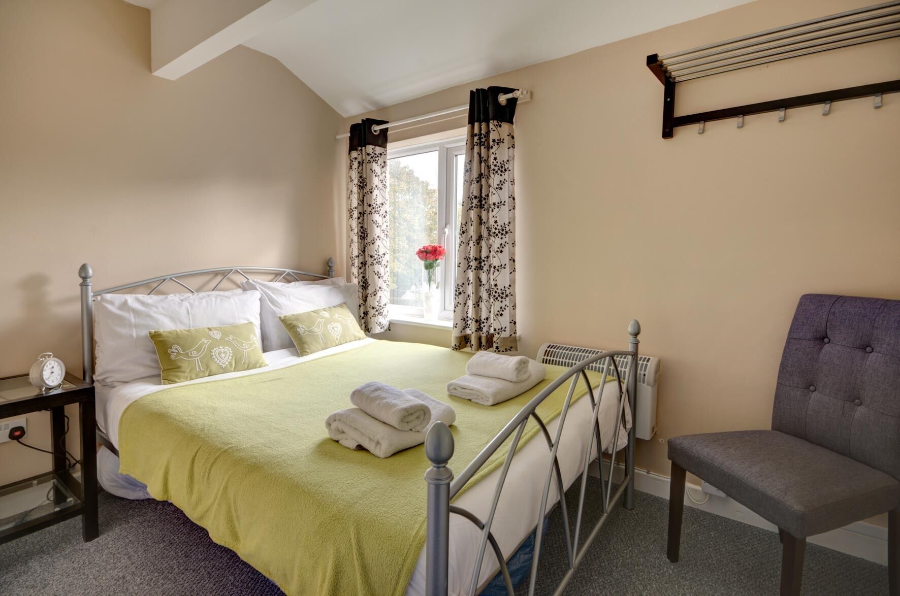 A neatly arranged bedroom with a bed, a chair, a bedside table with a clock, and a window with curtains.