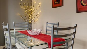 Un comedor con una mesa de vidrio, cuatro sillas de metal, un florero con luces y dos cuadros abstractos en la pared.