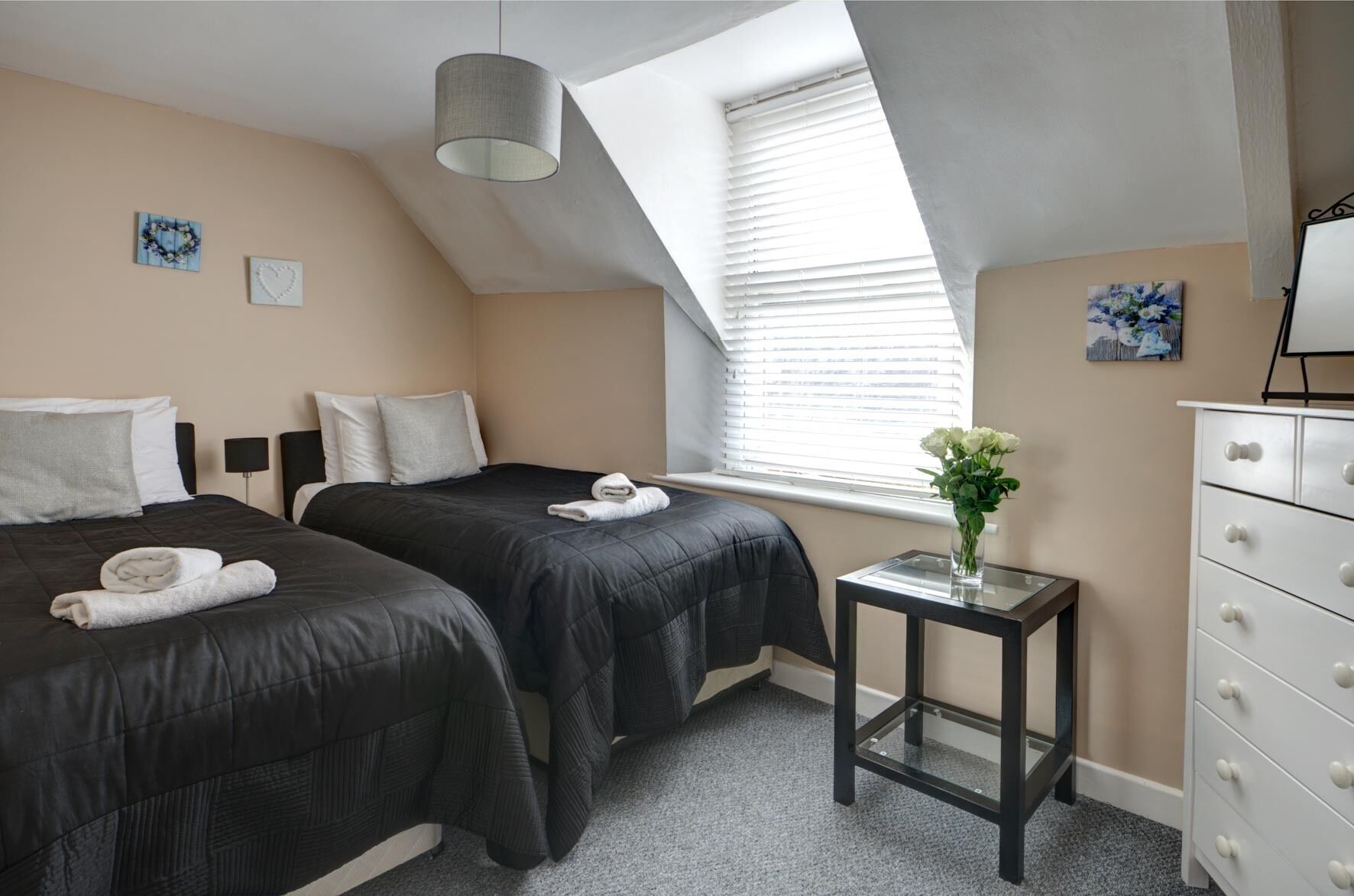 A bedroom with two beds, a small table, a dresser, and a window with blinds.