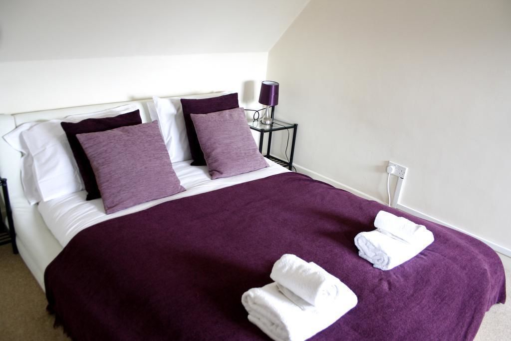 A neatly made bed with a purple bedspread and two pillows, a small bedside table with a lamp, and two folded towels on the bed.