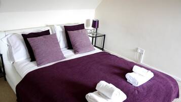 A neatly made bed with a purple bedspread and two pillows, a small bedside table with a lamp, and two folded towels on the bed.