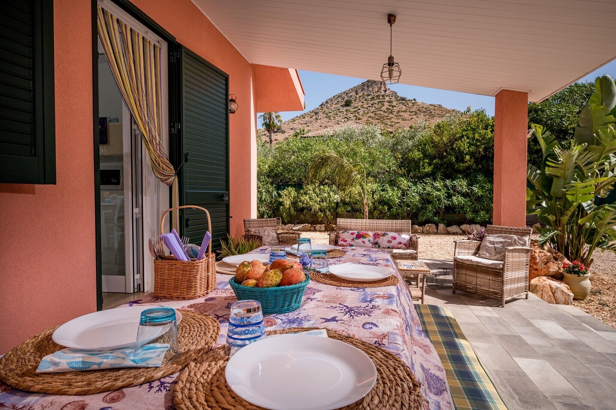 An outdoor dining area with a table set for two, wicker furniture, and a view of a mountain.