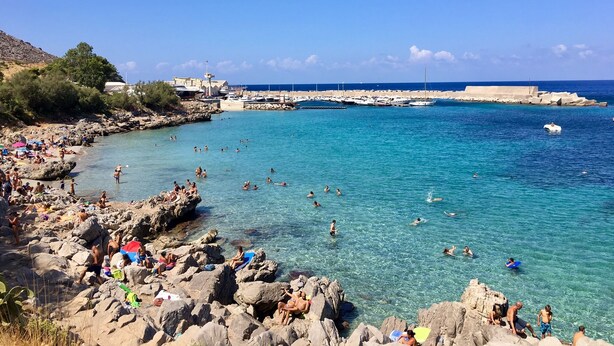 A rocky beach with clear blue water, people swimming and sunbathing, and boats docked in the background.