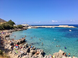 A rocky beach with clear blue water, people swimming and sunbathing, and boats docked in the background.