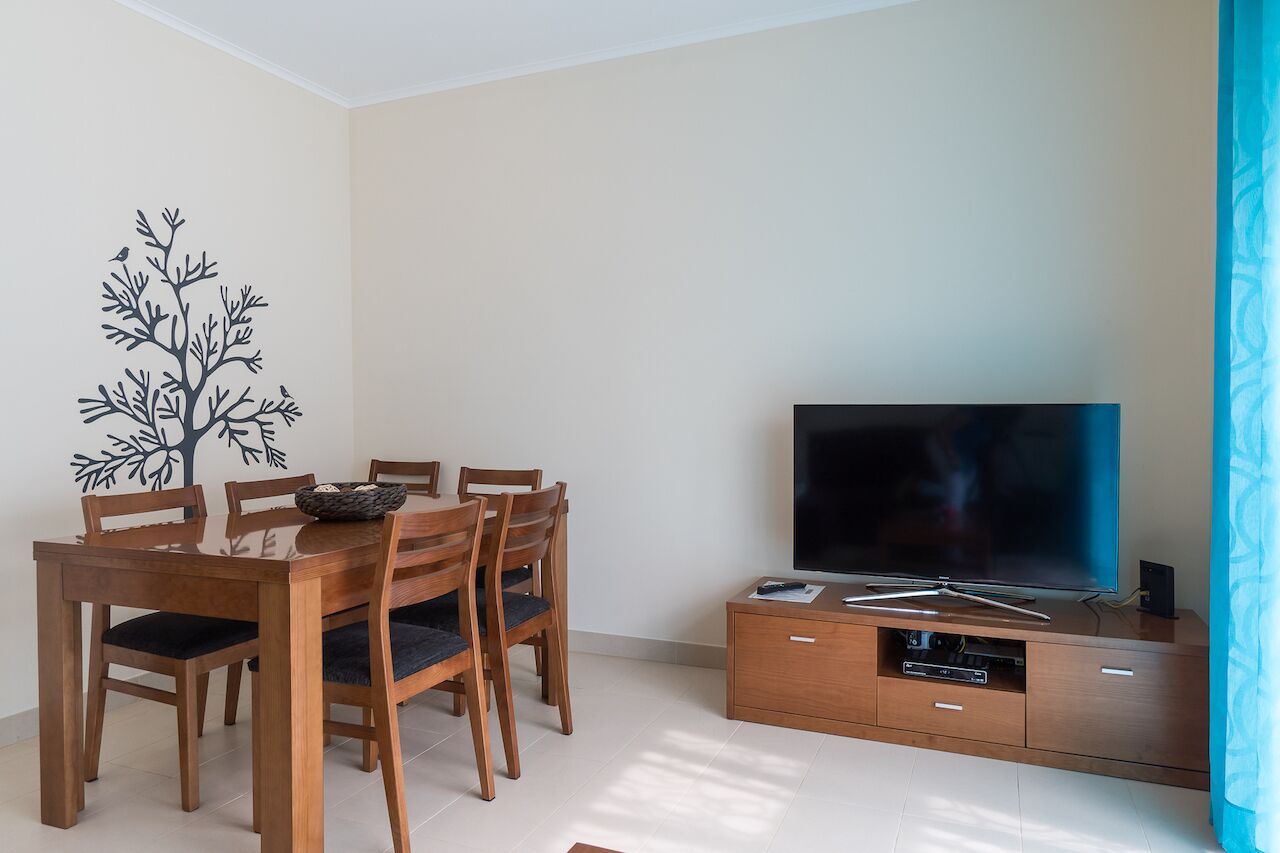 Un comedor con una mesa y sillas de madera, un televisor de pantalla plana sobre un mueble de madera, y un adhesivo mural con forma de árbol.