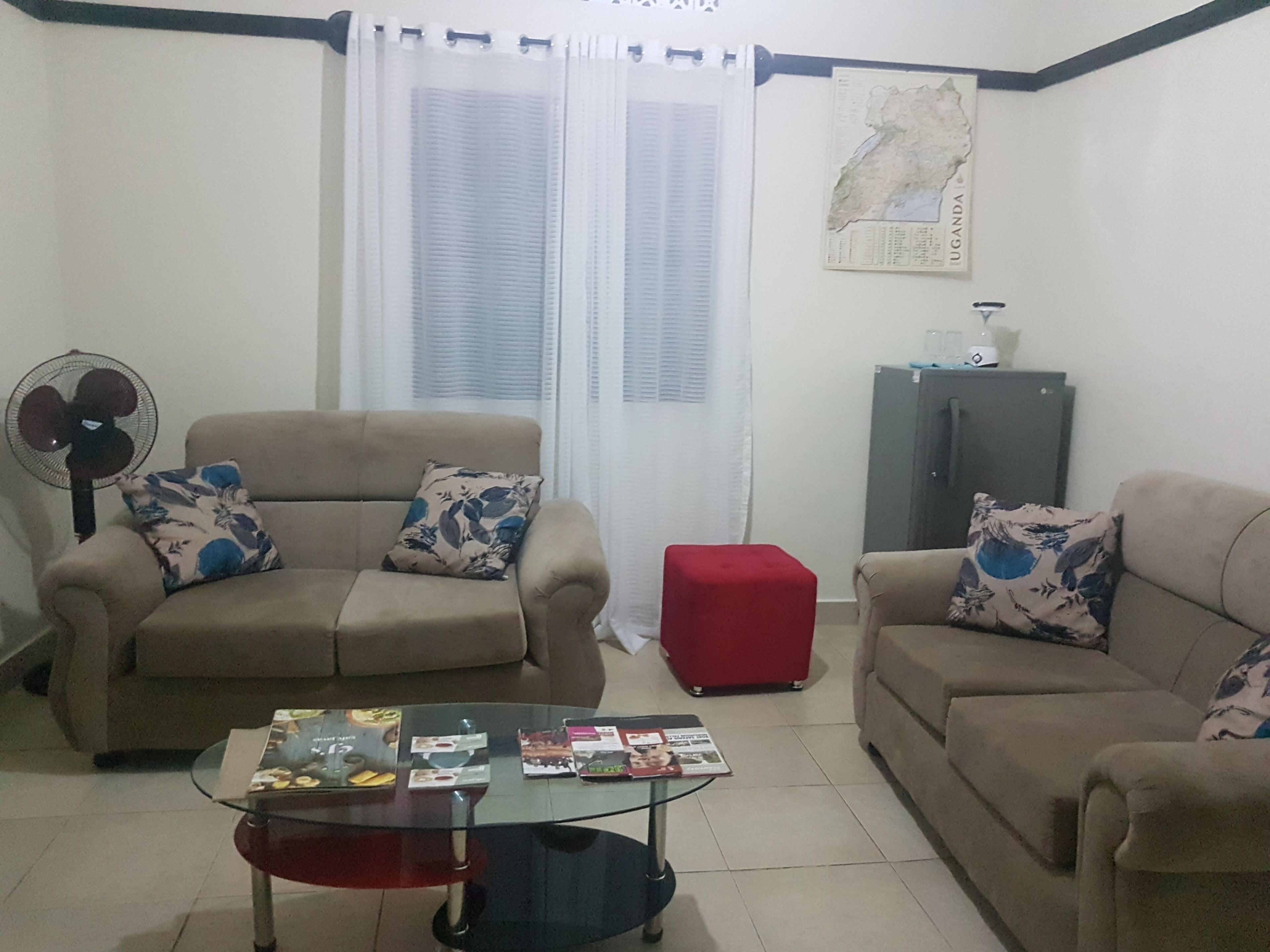 A living room with two beige sofas, a glass coffee table, a red ottoman, a fan, and a map on the wall.