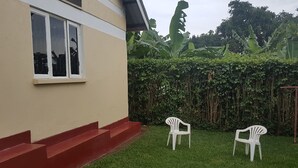 Una casa con fachada color beige y escalones rojos que conducen a una puerta blanca. Hay dos sillas de plástico blanco en el jardín delantero, cubierto de césped verde.