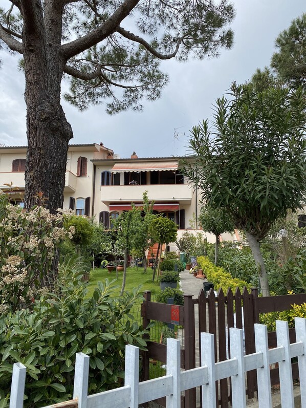 A two-story building with a garden in front, a white picket fence, and a large tree.