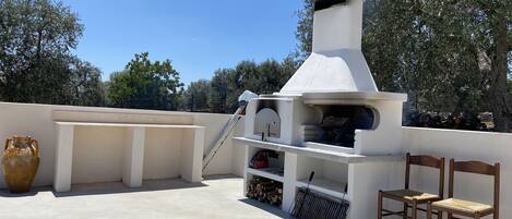 Un patio exterior con una parrilla blanca, dos sillas de madera, un jarrón grande y una pila de leña.