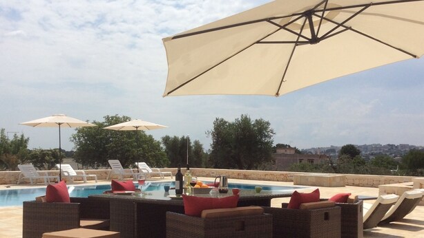 Zona junto a la piscina con mobiliario de mimbre y sombrillas, una piscina y vistas de la ciudad.