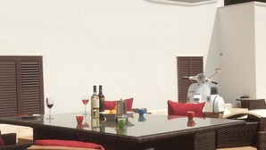 Un área de comedor al aire libre con mobiliario de mimbre, una mesa con copas de vino, botellas y una cafetera.