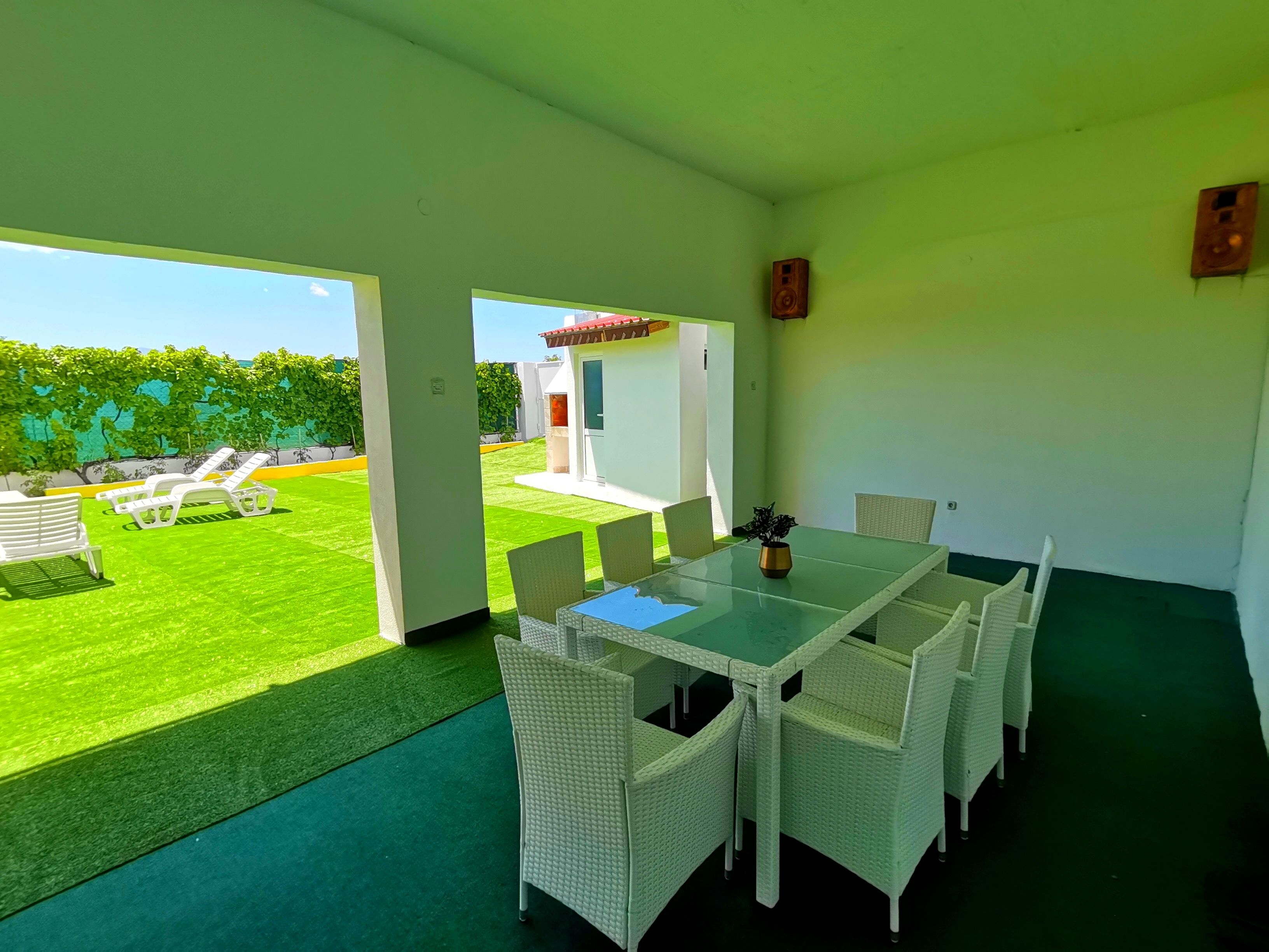 Ruime kamer met een glazen tafel en witte rieten stoelen, met uitzicht op een verzorgd gazon en een zitgedeelte buiten.