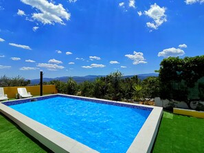 Ein rechteckiger Swimmingpool mit Fliesenrand, umgeben von grünem Kunstrasen, weißen Liegestühlen und Blumentöpfen.