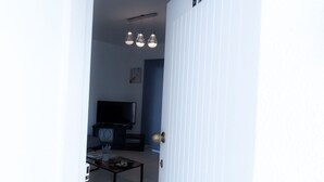 Une porte ouverte donne sur un salon où l’on aperçoit un téléviseur, une table basse et un luminaire fixé au mur.