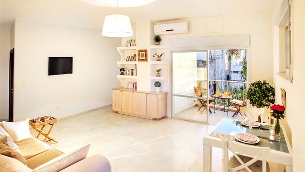 A modern living room with a sofa, a dining table with chairs, a bookshelf, and a sliding glass door leading to an outdoor balcony. - 2 BDR with Balcony ► Best Location: German Colony - Shelter in the building (Jerusalem)
