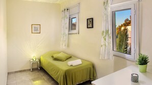 A small, well-lit bedroom with a single bed, a bedside table, a window with curtains, and a view of a building outside. - 2 BDR with Balcony ► Best Location: German Colony - Shelter in the building (Jerusalem)
