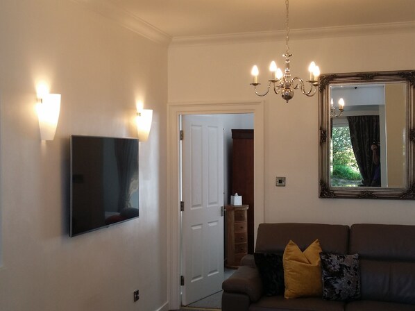 A living room with a flat-screen TV, a chandelier, a large mirror, and a brown sofa with cushions. - Dunperrogh Golfers paradise, ideal location 50 yards from 18th of Old Course (St Andrews)