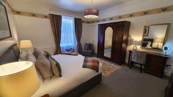 A hotel room with a large bed, a desk with a mirror, a chair, a wardrobe, and a window with curtains.