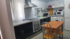 Cuina compacta amb fogó, forn, rentamics i microones. Hi ha una taula de dinar amb cadires i una finestra amb cortines.