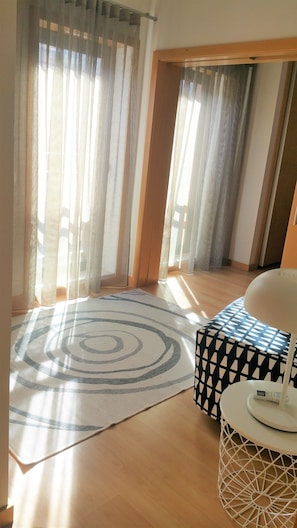 A room with a large window, a patterned rug, a geometric ottoman, and a white table with a device on it.