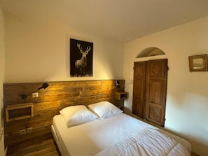 Une chambre à coucher avec une tête de lit en bois, un lit recouvert de draps blancs, un tableau représentant un cerf accroché au mur, une armoire en bois et une petite table de chevet avec une lampe.