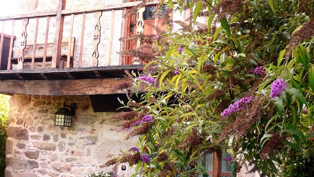 Un bâtiment rustique en pierre, doté d’un balcon en bois, de plantes en pot et d’un puits en pierre.