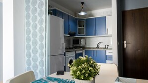 A modern kitchen with blue cabinets, a white refrigerator, a microwave, and a washing machine. There's a dining area with a table and chairs, and a potted plant on the table. - LOS NARANJOS APARTMENT.  (Matalascañas)