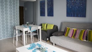 A modern living room with a grey sofa, colorful cushions, a white table with chairs, and abstract blue paintings on the wall. - LOS NARANJOS APARTMENT.  (Matalascañas)