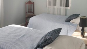 A bedroom with two beds, a chair, a window with sheer curtains, and a hanging light fixture.