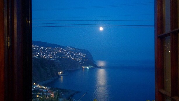 A night view from a window overlooking a coastal town with a visible moon and a body of water.