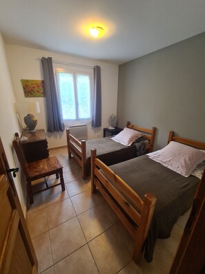 A room with two wooden beds, a small table, a chair, a radiator, a window with curtains, and a ceiling light.