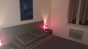Ein Schlafzimmer mit einem Bett, Kissen, einem Nachttisch mit roter Lampe, einem Wandbild und einem Fenster mit Vorhängen.