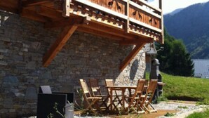 A wooden chalet with a stone wall, a balcony with wooden railings, and a table and chairs outside.