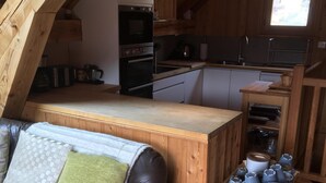 A loft-style living space with a kitchenette, a leather sofa, and a wooden dining table with a fruit bowl.
