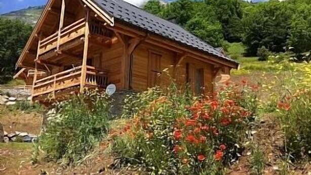 A wooden cabin with a balcony, surrounded by a garden with various plants and flowers.