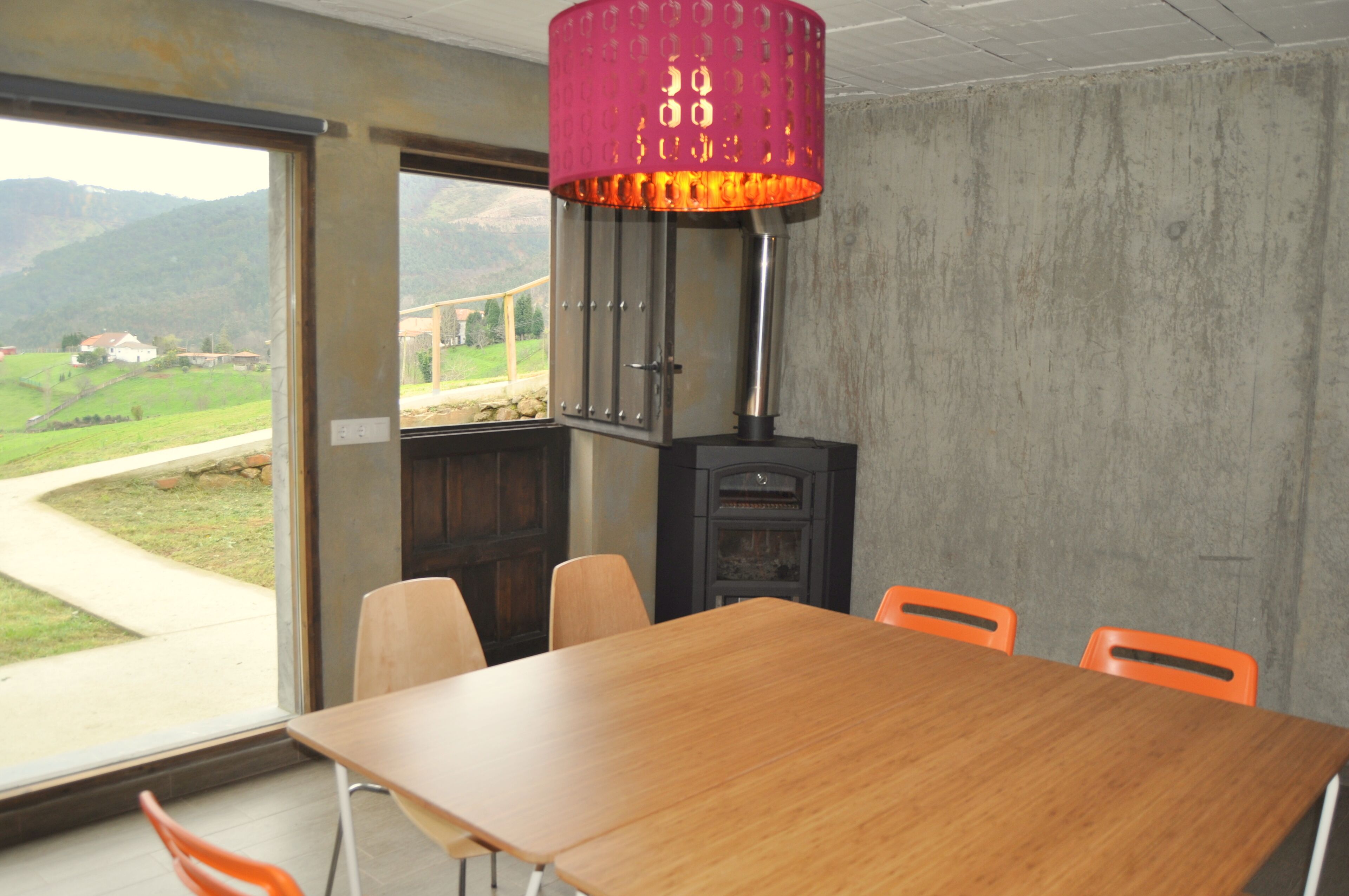 Un salón de conferencias con una mesa de madera, sillas anaranjadas, una lámpara roja y vistas a un paisaje verde a través de un ventanal grande.