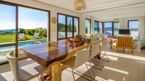 Un vaste espace de vie comprenant une grande table à manger en bois, des chaises blanches, un canapé et un téléviseur. De grandes fenêtres offrent une vue sur une piscine et sur la verdure environnante.