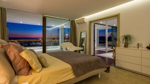 Une chambre d’hôtel moderne avec un grand lit, une vue sur la ville au crépuscule, un balcon et une commode blanche.