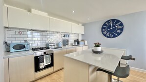 A modern kitchen with white cabinets, a blue microwave, a stove, and a sink. There's a large blue clock on the wall and a small potted plant on the counter. - A CONTEMPORARY BEACHSIDE APARTMENT ON HEADLAND ROAD FISTRAL 5 MINUTE FROM BEACH  (Cornwall)