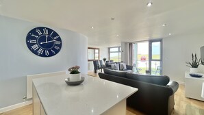 A modern living room with a large wall clock, a dining area, and a view of the outdoors. - A CONTEMPORARY BEACHSIDE APARTMENT ON HEADLAND ROAD FISTRAL 5 MINUTE FROM BEACH  (Cornwall)